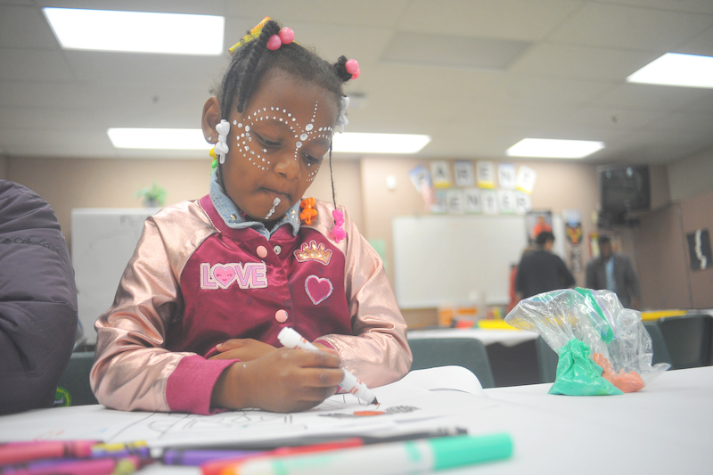 Black girl with rhinestones on her face colors with marker in a classroom.