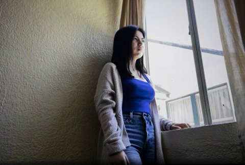 Young woman in sweater, blue shirt and jeans standing next to a window.