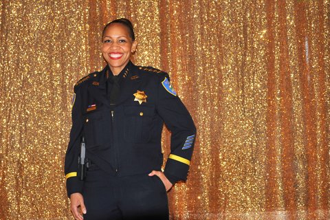 A smiling Black and Latina woman in police uniform in front of sparkly gold curtain.