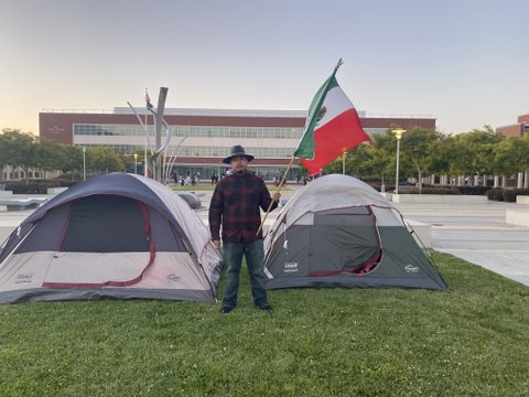 Man holding Mexican flag by two tents on grass with building in background