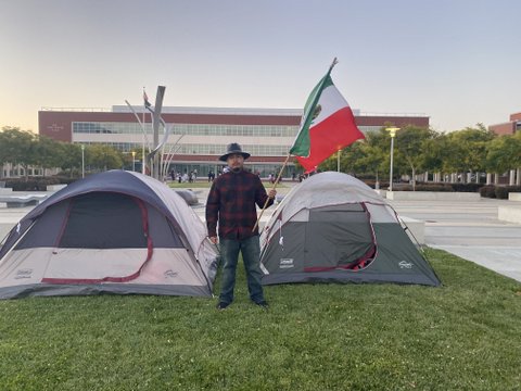 Man holding Mexican flag by two tents on grass with building in background.