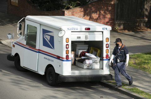 Postal carrier in mask and gloves next to the back of his open mail truck.