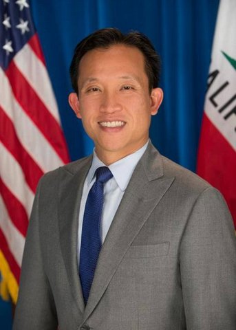 Asian man in gray suit and blue tie with U.S. and California flags partly visible behind him.