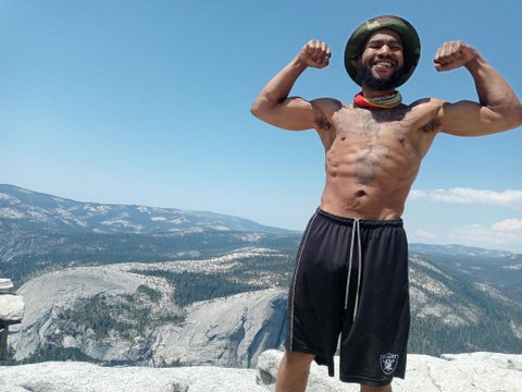 A shirtless Black man in hat and shorts flexes his biceps standing on top of Half Dome Mountain.