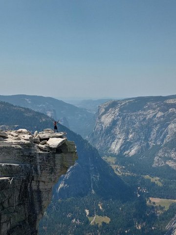 Wide shot of Yosemite with man standing atop Half Dome Mountain with arms raised triumphantly.