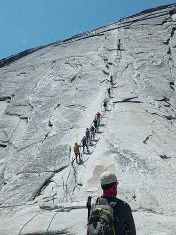 View from behind of man standing at the base of a sheer rock wall that people are using cables to ascend.