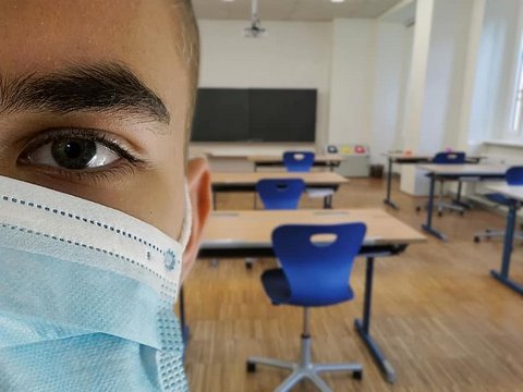 Close up of right side of face of young man wearing medical face mask with empty classroom in background