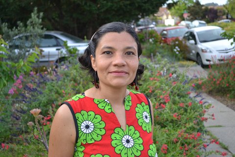 A Latina woman in a red dress with green flowers standing in front of a sidewalk garden.