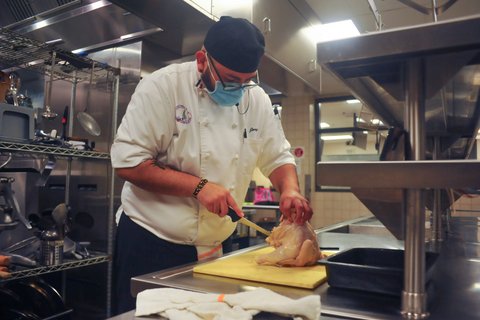 Man in white chef's coat, black cap, blue mask and glasses cuts a raw chicken.