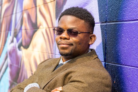 A Black man wearing a suit and tinted glasses standing with his arms folded against a wall painted purple with a mural.