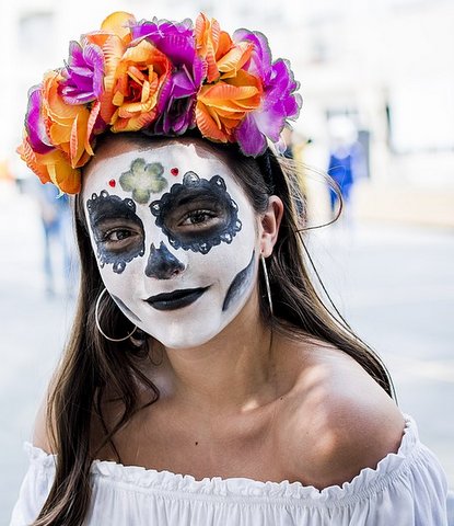 A young woman wearing skull face makeup and a headdress of orange and purple flowers.