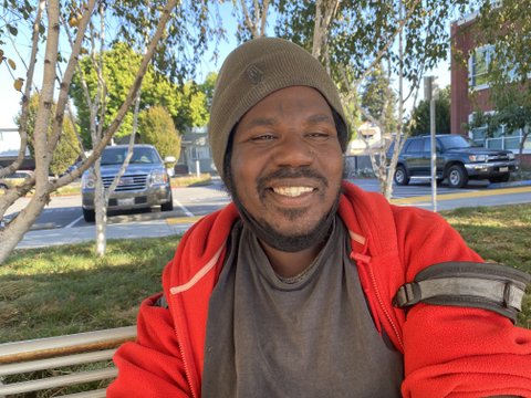 A smiling Black man wearing a beanie and red jacket with his face mask down around his chin.