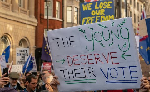 Scene from a protest march with person holding sign in foreground that says "The young deserve their vote." Sign in the background says "We lose our freedom."