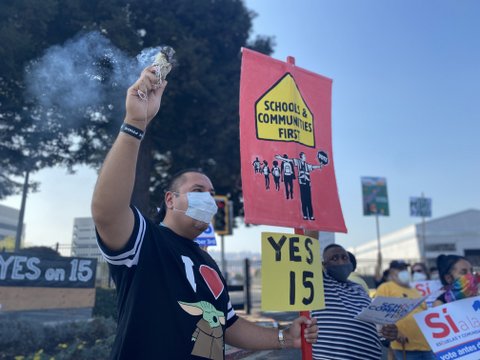 A man in medical face mask holds one sign that says "yes 15" and one that says "schools and communities first" with a drawing of kids and a crossing guard holding a "stop" sign that says "vote." The man is wearing a black shirt with the letter "I" and drawings of a heart and Baby Yoda.