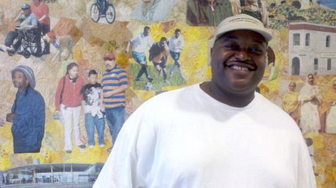 A smiling Black man wearing a cap and white T-shirt in front of a mural showing different types of people
