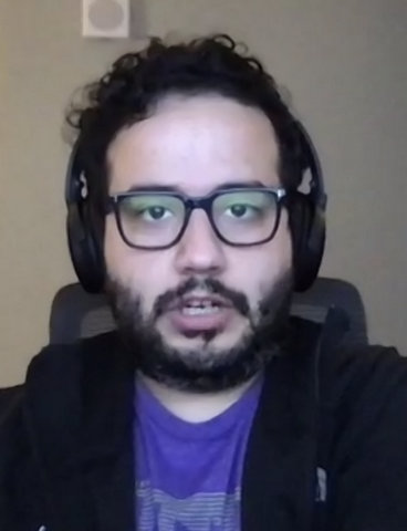 A Latino man with beard, black-framed glasses, headphones, dark jacket and purple shirt.