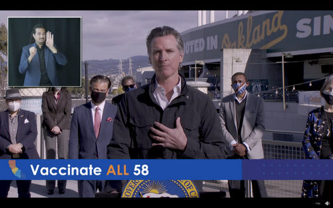 Gavin Newsom and masked people outside Oakland Coliseum. Chyron says "vaccinate all 58."