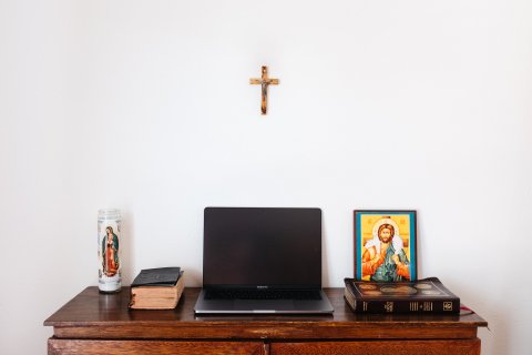 Laptop on table with bibles, Virgin Mary candle and drawing of Jesus. A crucifix is on the wall behind.