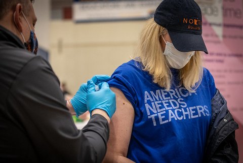 Blonde woman wearing black baseball cap, white face mask and blue T-shirt that says "America needs teachers" getting vaccinated.