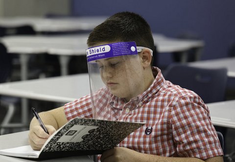 Boy in classroom wearing a face shield and writing in a composition notebook