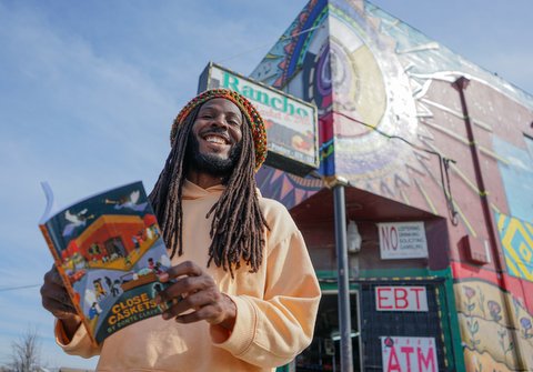 Looking up at smiling Black man with beard and dreds, holding book in front of market.