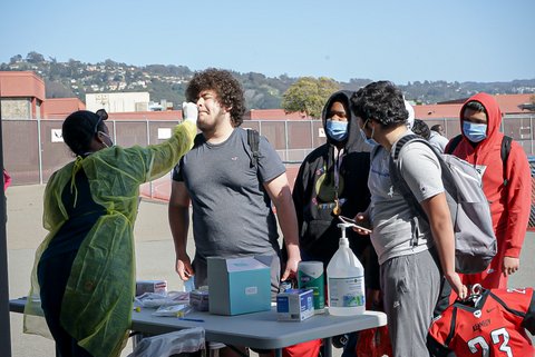 Medical worker administers nasal swab while others in masks wait in line.