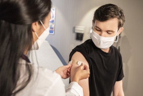 Woman puts bandaid on young man's arm. Both are wearing face masks