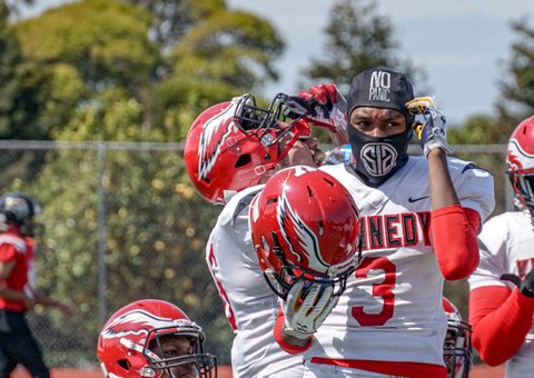 Football player wearing head covering that says "no panic" and a mask holds his helmet.