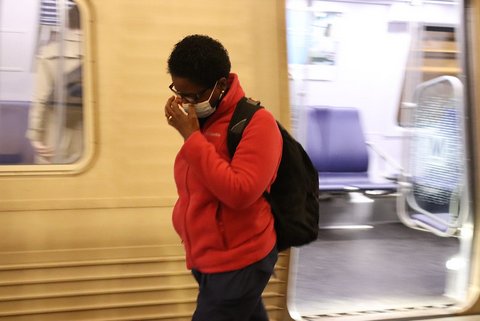 Black woman in mask, glasses, backpack and red jacket walks in front of a subway car with her hand to her face.