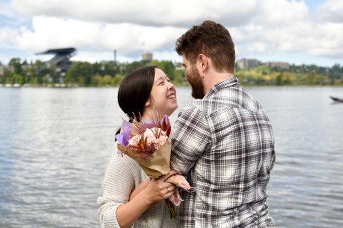 Man and woman standing together, facing each other, in front of a lake.