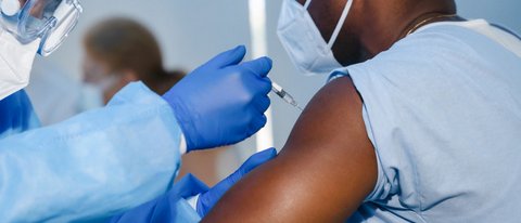 Closeup of a Black man getting a shot in his arm. He is wearing a mask, and the medical worker is in protective gear.