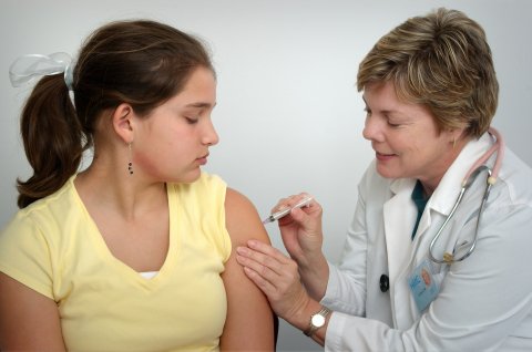 Adolescent girl in yellow shirt and ponytail looks at her left arm while a nurse in white lab coat gives her a vaccine shot.