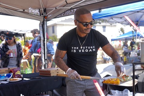 A Black man with tattoos on his arms, a cross necklace, dark sunglasses and a black T-shirt that says "The future is vegan" making pizza in an event booth.