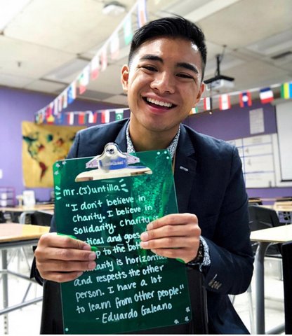A smiling young Latino man in a classroom, wearing a blue suit and holding a green clipboard