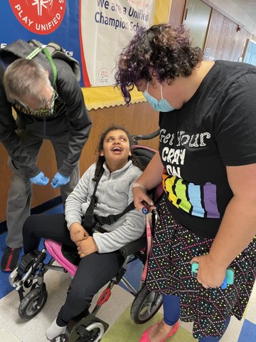 A man and woman in masks looking down at a girl in a wheelchair who is looking up at the woman.