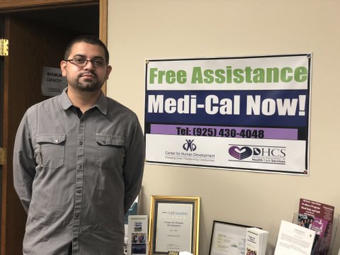 Latino man with close cut hair and beard, glasses and gray button-down shirt next to a table with pamphlets and other written material and a sign that says "Free assistance Medi-Cal now!"