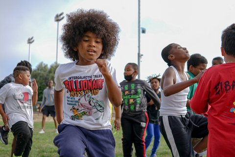 A boy with an afro and a white T-shirt that says "intergalactic tales billionaire boys club" runs on a field among other boys.