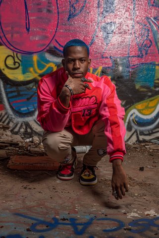 A Black man with short, blue hair and a pink and red jacket kneeling in front of a wall painted with graffiti looking straight ahead.