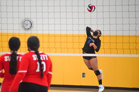 Girl in a black top, shorts and mask jumps to serve a volleyball. Opponents in red are seen in the foreground.