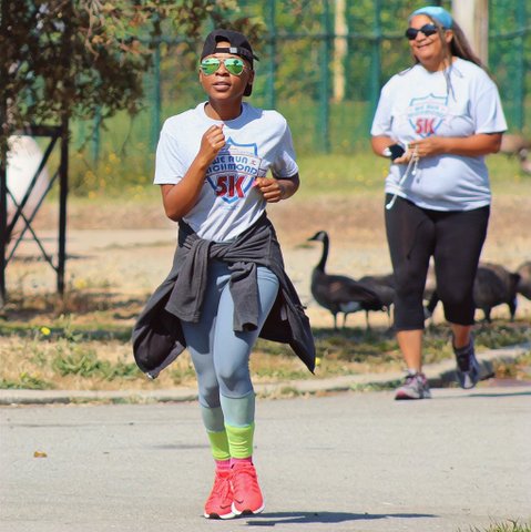 A Black person running in backwards baseball cap, green aviator sunglasses, We Run Richmond 5K T-shirt, jacket tied around their waist, blue pants, green socks and reddish shoes