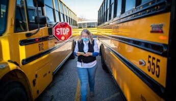 An older white woman in medical mask walks between two school buses, one of which has its stop sign out