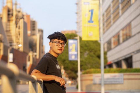 A young Asian man with glasses standing on an out-of-focus college campus.