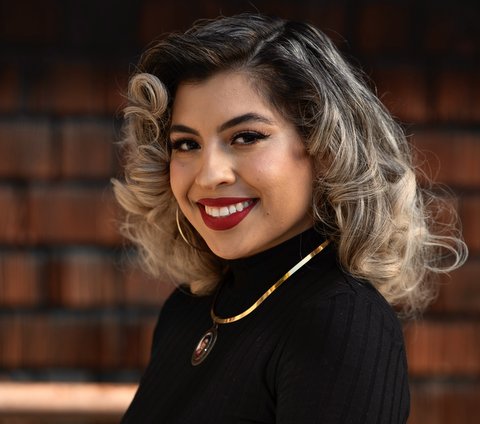 A smiling Latina young woman with curly hair partially dyed blond, red lipstick, black sweater and gold necklace.