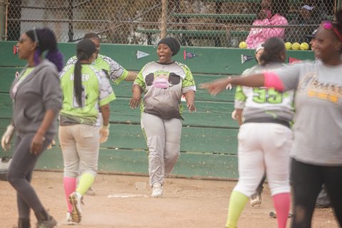 A group of Black women near home plate during a softball game. Woman in the center has a joyful expression.