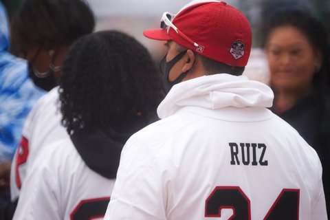 View from behind of a man in a red cap, mask and white hoodie that says 'Ruiz 24" on the back.