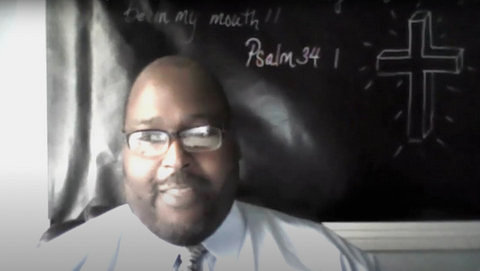 A Black man in front of a chalkboard with a drawing of a cross, the words "Psalm 34:1" and part of the psalm visible.