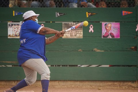 Side view of a Black woman in white cap, gray pants and blue mask and jersey hitting a yellow softball
