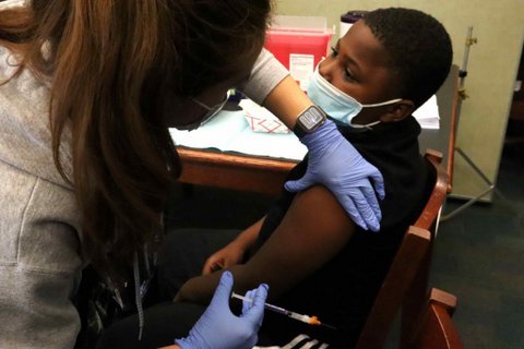 A young Black boy looks toward a medical worker as she prepares to give him a shot.