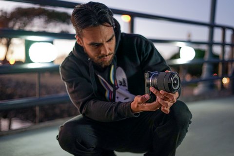 A young Latino man crouching on a bridge looking at a camera he is pointing to his left.