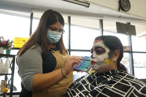 A young woman wearing a medical mask, glasses and an apron applies makeup to a woman made up for Day of the Dead.